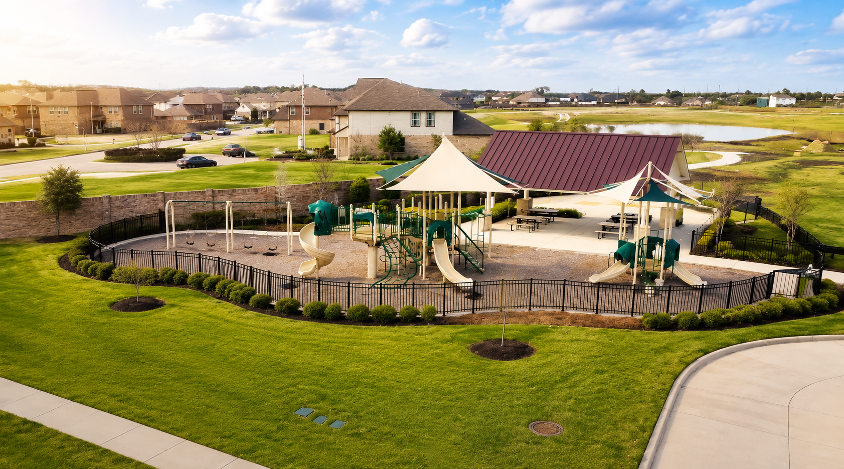  Aerial view of a modern community playground featuring shaded play structures, slides, swings, and a pavilion surrounded by manicured lawns, walking paths, and a suburban neighborhood under a sunny sky.