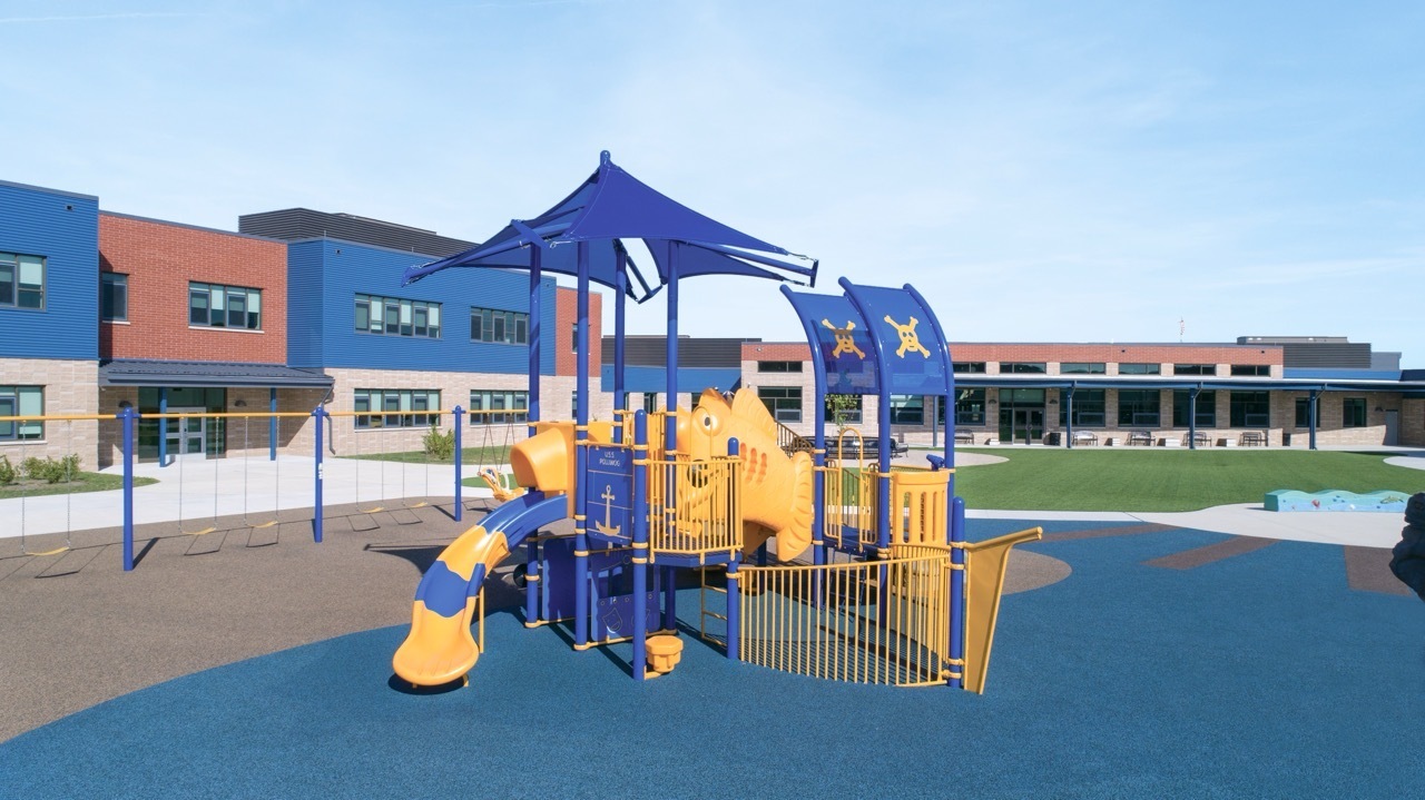 Colorful blue and yellow elementary school playground featuring slides, climbing structures, swings, shade canopies, and rubber safety surfacing outside a modern school building.