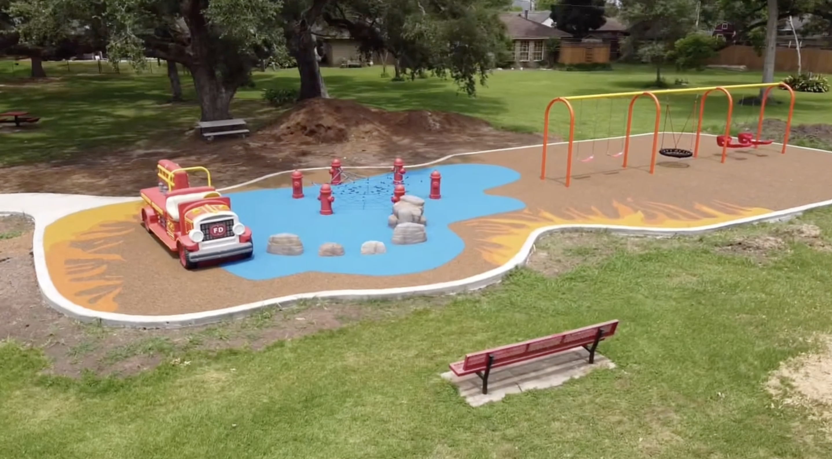 A firefighter-themed playground featuring a red fire truck play structure, fire hydrant obstacles, climbing elements, colorful surfacing designed like water and flames, and a row of swings.