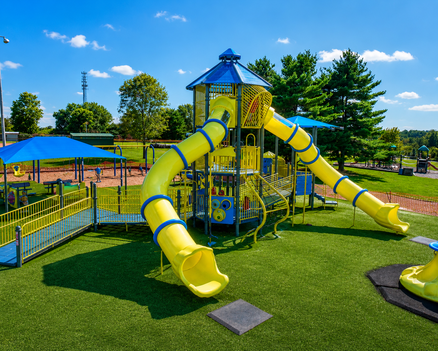 A colorful commercial playground with bright yellow tube slides, climbing structures, and blue shade canopies in a sunny community park, surrounded by green trees, open grassy areas, and additional play equipment in the background.