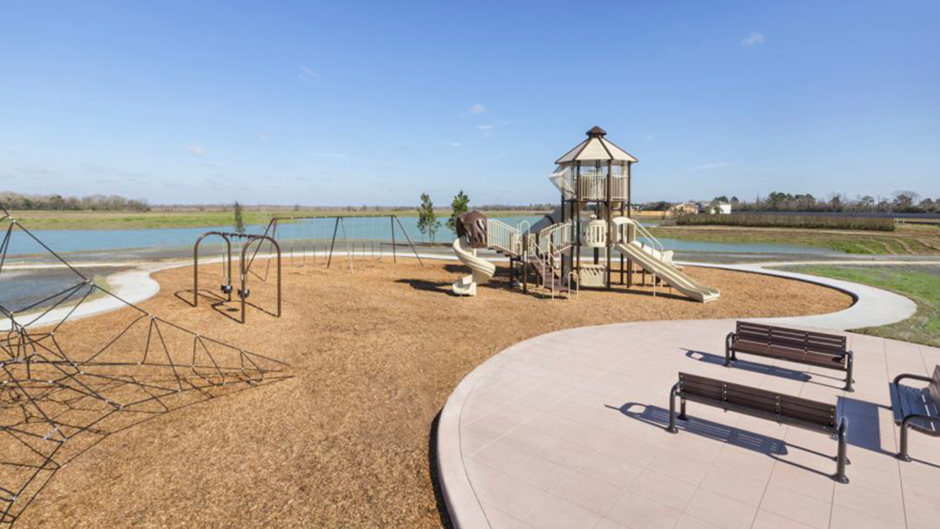 Community playground featuring slides, swings, climbing structures, and seating areas beside a scenic waterfront, with open skies and walking paths surrounding the recreation space.