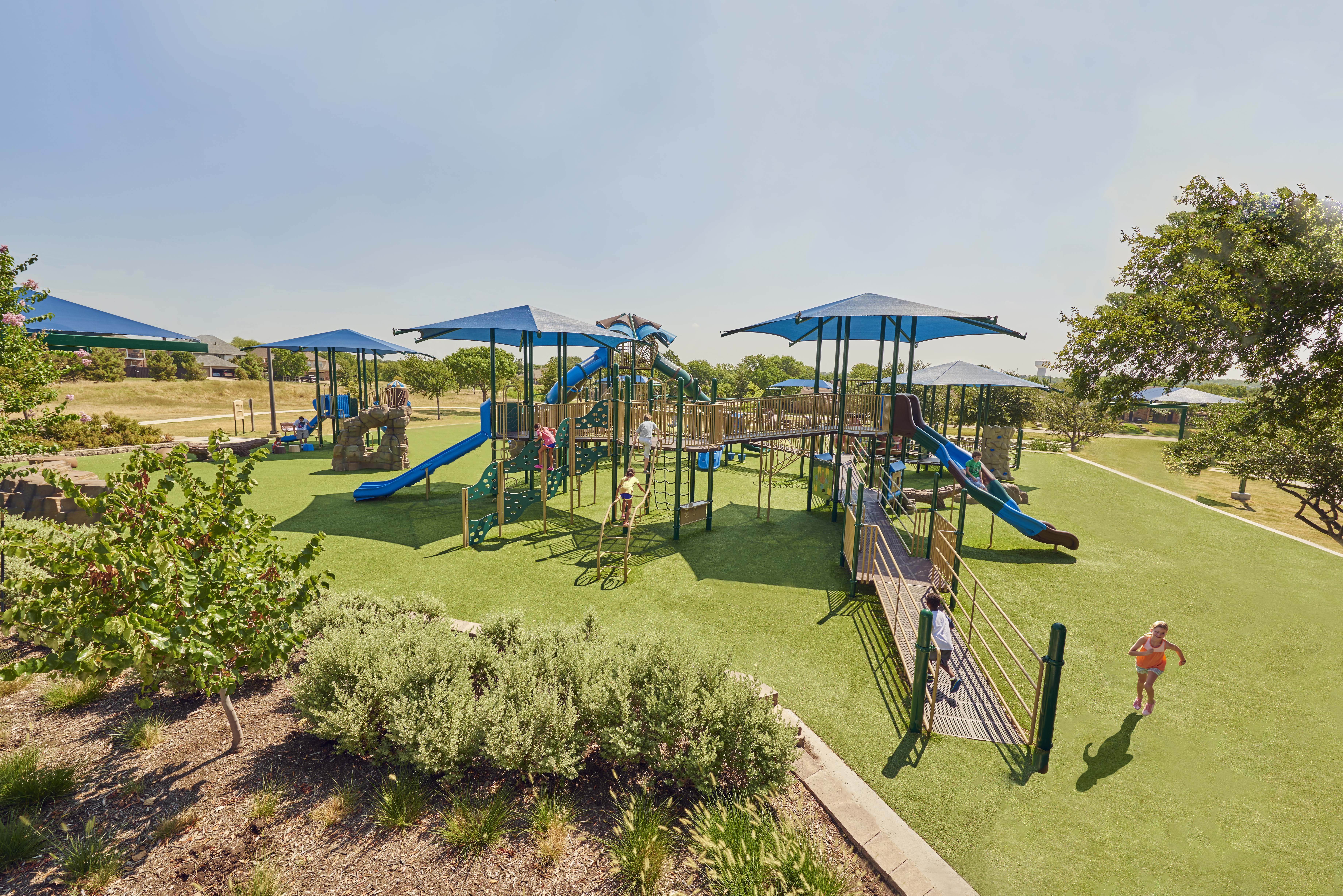 Large commercial playground with blue shade structures, slides, climbing equipment, accessible ramps, and artificial turf surrounded by trees and landscaping.